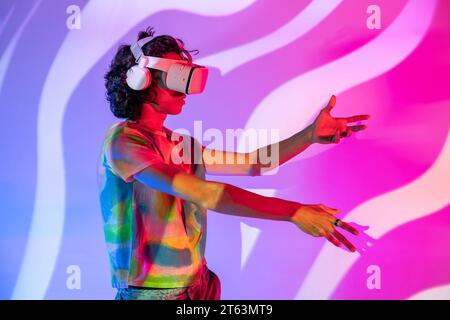 Young man wearing a tie-dye shirt and VR headset reaches out, immersed in the digital world, complemented by a wavy pink and purple background Stock Photo