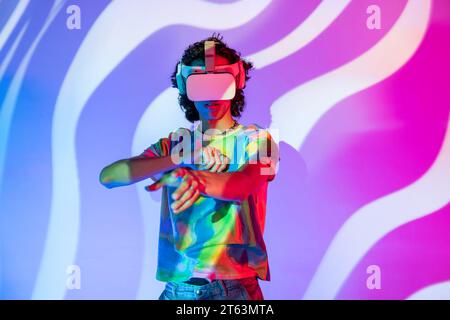 A man in a vibrant tie-dye shirt interacts with a virtual world through his VR headset, set against a swirling purple and blue backdrop Stock Photo