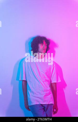 A contemplative young man with curly hair, clad in a plain white shirt, stands against a gradient background transitioning from purple to blue Stock Photo