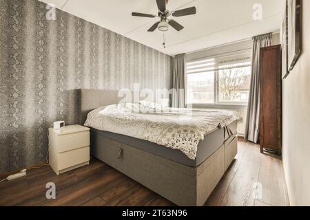 Ceiling fan over comfortable white bed and gray wallpaper on wall by large bright window in spacious bedroom at home Stock Photo