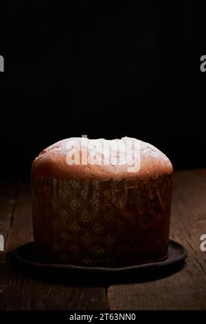 A freshly baked panettone sits on a rustic wooden table, highlighted against a dark background, creating an intimate and inviting scene Stock Photo