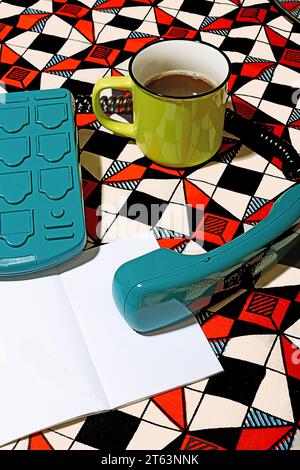 High angle of turquoise vintage telephone alongside a green mug filled with coffee, placed on a striking geometric-patterned backdrop near blank white Stock Photo