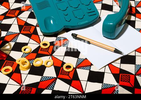 High angle of vintage turquoise telephone with its coiled cord extending rests on a vibrant geometric-patterned background near blank white paper shar Stock Photo
