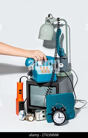 Hand holding a vintage telephone in a curated collection of vintage technology items against a white backdrop, featuring an old television, alarm cloc Stock Photo
