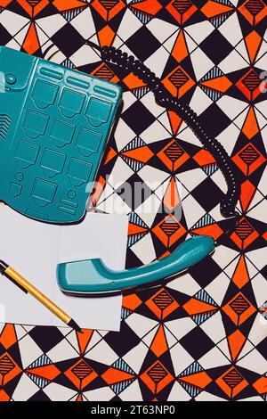 Top view of vintage turquoise telephone with its coiled cord extending rests on a vibrant geometric-patterned background near blank white paper and a Stock Photo