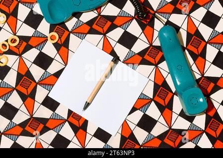 Top view of vintage turquoise telephone with its coiled cord extending rests on a vibrant geometric-patterned background near blank white paper sharpe Stock Photo