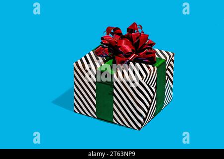 striped black and white gift box adorned with a shiny red bow and a green ribbon placed against a vibrant turquoise background Stock Photo