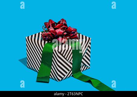 striped black and white gift box adorned with a shiny red bow and a green ribbon placed against a vibrant turquoise background Stock Photo