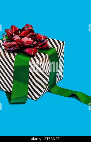 striped black and white gift box adorned with a shiny red bow and a green ribbon placed against a vibrant turquoise background Stock Photo