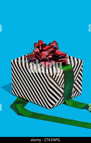 striped black and white gift box adorned with a shiny red bow and a green ribbon placed against a vibrant turquoise background Stock Photo