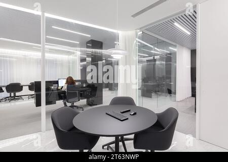 Spacious modern office layout illuminated by bright overhead lights, showcasing workstations with computers and chairs, separated by glass partitions, Stock Photo