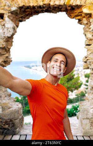 Joyful man with hat in an orange shirt taking a selfie with a coastal view of Barcelona city through a stone arch behind him. Stock Photo