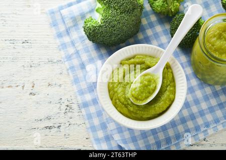 Green broccoli baby food in white bowl and jar on table. Green baby food. Child first feeding concept. Baby Natural Food. Production and menu of baby Stock Photo