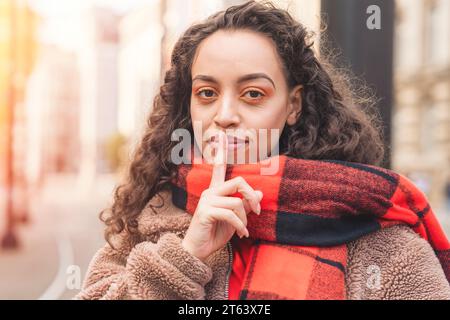 an ethnic curly-haired woman in a jacket and an orange jacket holding her index finger to her lips. Shushing concept Stock Photo