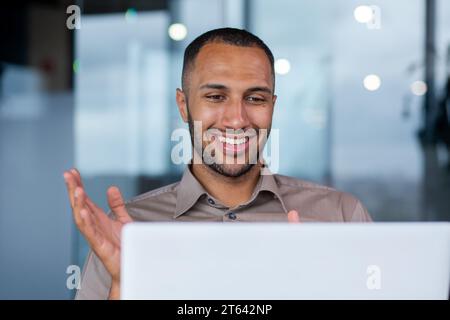 Close-up of a happy successful hispanic businessman making a video call from a laptop in a modern office, a young programmer is happy about a successful job, smiling at the computer screen. Stock Photo