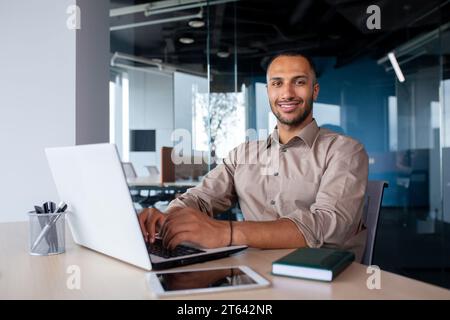 Attractive african american man sitting at desk in front of laptop in office, programmer working in modern environment, tapping on keyboard and smiling at camera. Stock Photo