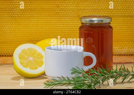 Jar of honey, small white cup, rosemary sprig and a lemon and a half on wood with a real honeycomb in the background Stock Photo