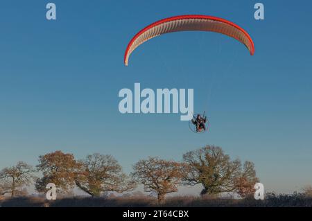 Para-glider awith large orange sail against a clear blue sky over the top of winter trees Stock Photo
