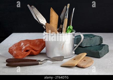 Modeling tools from plasticine and clay in a metal mug and pieces of clay and plasticine on a white concrete table on a black background Stock Photo