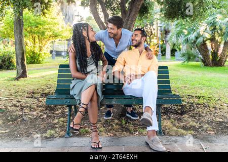 Three cheerful young adults, a diverse group, laughing and enjoying a sunny day on a park bench. Stock Photo
