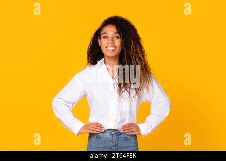 Black millennial lady standing holding hands on hips, yellow backdrop Stock Photo