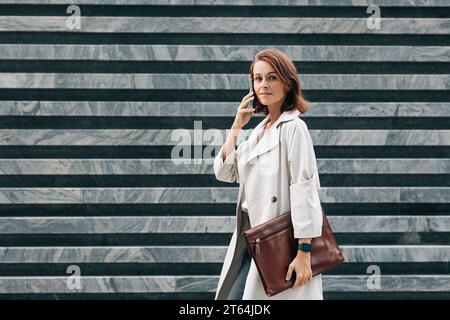 Side view of stylish middle-aged businesswoman in coat. Middle-aged female talking on mobile phone while walking. Stock Photo