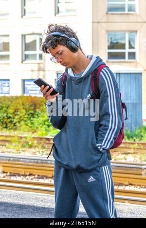 Young man wearing Adidas sportswear with smartphone and BOSE headset waiting train - Tours, Indre-et-Loire (37), France. Stock Photo