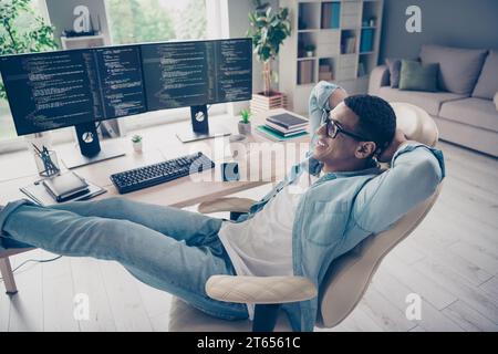 Full size photo of young guy relaxing put his legs technical support employee pause when code works on monitors indoors office background Stock Photo
