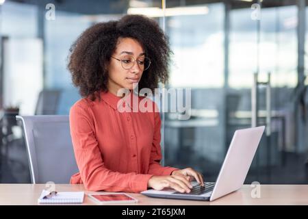 Businesswoman with laptop at workplace inside office, confident serious thinking woman typing on computer keyboard, female worker in red shirt with curly hair, focused and concentrated at work. Stock Photo