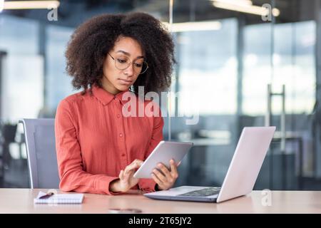 Serious confident woman working with tablet computer inside office, business woman thinking sitting at desk with laptop, business woman in shirt and curly hair. Stock Photo
