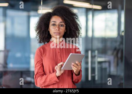 Serious confident woman in red shirt inside office with tablet computer in hands, looking at camera in concentration and thinking, businesswoman at workplace inside office, programmer developer. Stock Photo