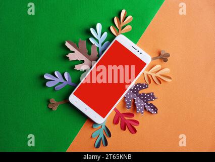 A white mobile phone with empty red blank space for text in the middle lies on a half green and orange background surrounded with paper leaves of red, Stock Photo