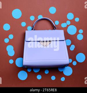 Trendy lavender leather female top-handle bag with black trim, metal accent, and feet isolated on a cartoonish brown paper background with blue circle Stock Photo