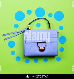 Fashionable women's blue leather handbag with flap, gold hardware, and top handle isolated on a fun green paper background with cut-out blue circles. Stock Photo