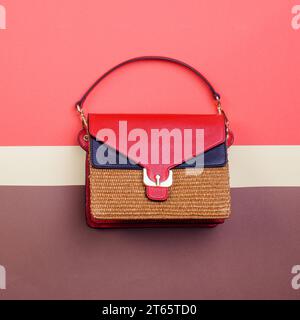 Modern women's woven handbag with red and black leather flap, gold hardware, and top handle isolated on a creative multicolor background with copy spa Stock Photo