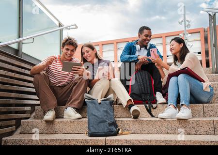 Group of multiracial high school students talking on a staircase at university campus, using a tablet app and smartphone to do homework project after class. Friendly teenage people studying together. High quality photo Stock Photo