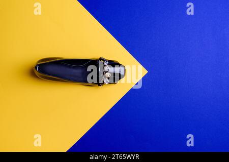 Bold woman's black patent leather shoe decorated with crystals on the front on a contrasting yellow-blue background with pointing on copy space. Creat Stock Photo