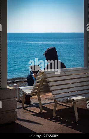 Guy in a hoodie sitting on a bench while working remotely on a laptop at Prom des Anglais, Nice, with the sea in the background; Remote work wallpaper Stock Photo