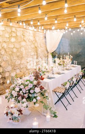 Bouquets of flowers and lit candles stand on a festive table on the terrace in the light of garlands Stock Photo