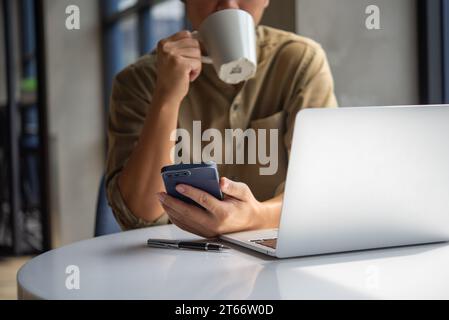 Man working on laptop and using phone while enjoying a refreshing drink coffee in a modern office. Business Digital technology communication and netwo Stock Photo