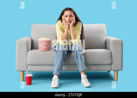 Young woman with popcorn watching movie on sofa against blue background Stock Photo
