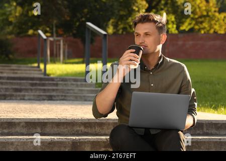 Handsome man with laptop drinking coffee on stairs outdoors. Space for text Stock Photo