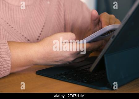 Female using phone looking at laptop or digital tablet device. Unrecognizable woman sitting at table in a cafe and use online banking with smartphone. Stock Photo