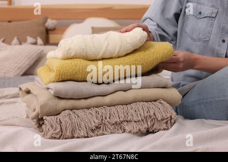 Woman with stack of different folded blankets and clothes on bed in room, closeup. Home textile Stock Photo