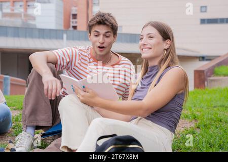 Two students on campus sitting surprised reviewing notes for exam outdoors. Stock Photo