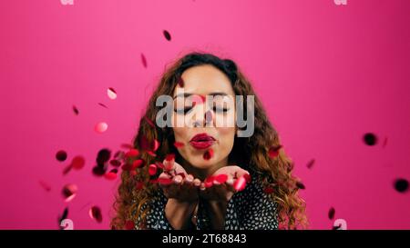 Beautiful young woman blows red confetti from palms of hand, Valentines party Stock Photo