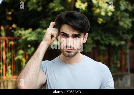Confused man scratching head in park on sunny day Stock Photo