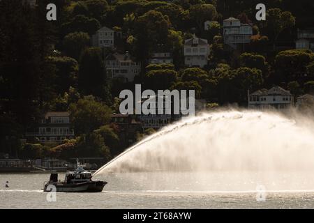 Tiburon, California, USA - 04, 25, 2023: Black and white boat floating on rippling sea with water splashes near city with residential buildings agains Stock Photo
