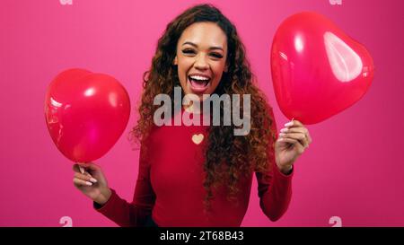 Cute young woman dances with heart shaped balloons, waves in air, studio Stock Photo