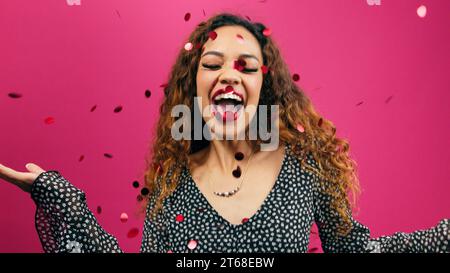 Beautiful young woman dances while red confetti glitter rains down, pink studio Stock Photo